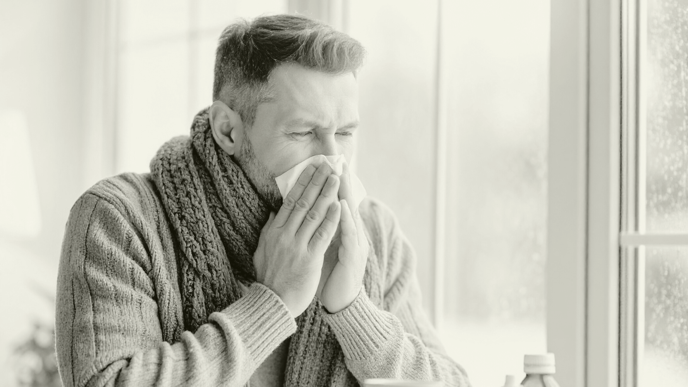Man with a cold sitting by a window, blowing his nose, representing winter illnesses and seasonal pressures in Primary Care.