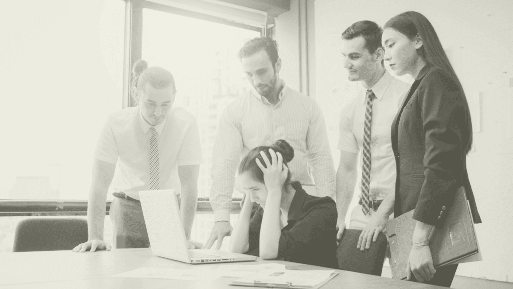 A stressed healthcare manager surrounded by colleagues while planning staffing and rotas on a laptop, representing the impact of winter pressures, financial year demands and holiday cover in primary care.