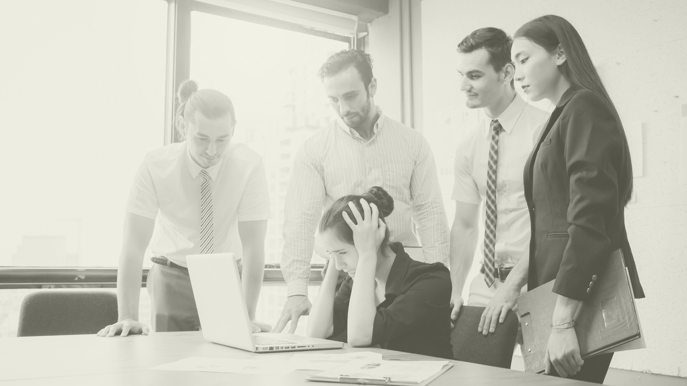 A stressed healthcare manager surrounded by colleagues while planning staffing and rotas on a laptop, representing the impact of winter pressures, financial year demands and holiday cover in primary care.